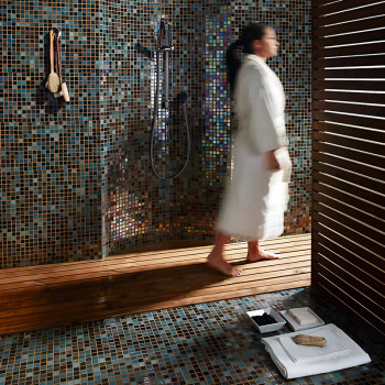 Person walks on wooden floor in mosaic-tiled bathroom, with a showerhead and hanging brushes on the wall.
