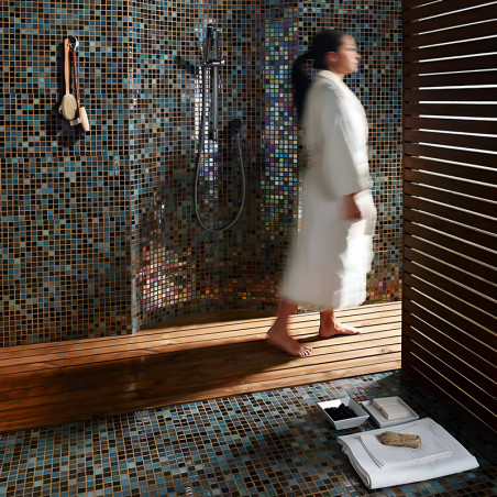 Person walks on wooden floor in mosaic-tiled bathroom, with a showerhead and hanging brushes on the wall.