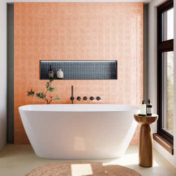 In een zonovergoten badkamer rust een wit vrijstaand bad tegen een perzikkleurige tegelwand met een verzonken zwarte plank met d