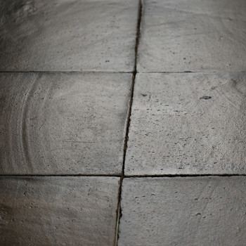 Square terracotta tiles lie adjacent, subtly textured with circular patterns, forming a weathered floor in dim lighting.