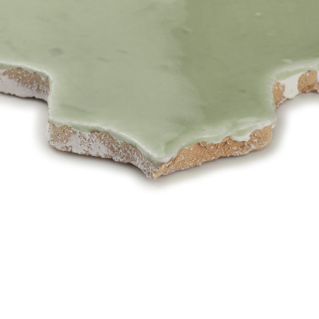 Un carreau en terre cuite vernissée verte posé sur une surface blanche, présentant des bords rugueux et inégaux et une finition 