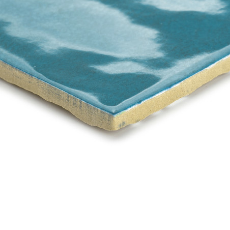 Un carreau de céramique présente un motif bleu ondulé et brillant sur sa surface, avec un bord beige clair et rugueux, conférant
