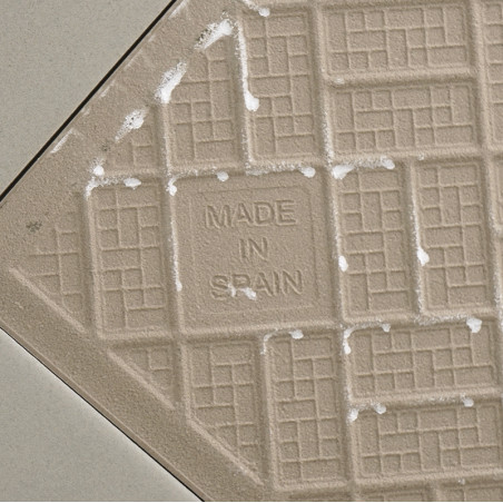 Een beige keramische tegel heeft een rasterpatroon met in het midden de tekst `MADE IN SPAIN`, omgeven door lichte, diagonale vo