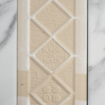 Eine beige, strukturierte Fliese zeigt geprägte Rautenformen mit dem Text „MADE IN SWEDEN“ in der Mitte. Es liegt vor einem Hint