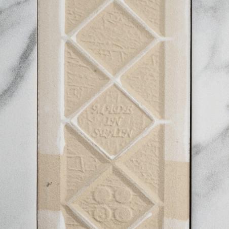Eine beige, strukturierte Fliese zeigt geprägte Rautenformen mit dem Text „MADE IN SWEDEN“ in der Mitte. Es liegt vor einem Hint