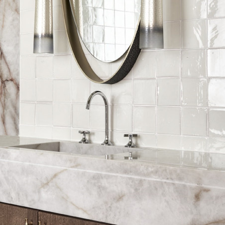 White terracotta tiles line a bathroom wall, reflecting light around a marble countertop with a modern faucet fixture.