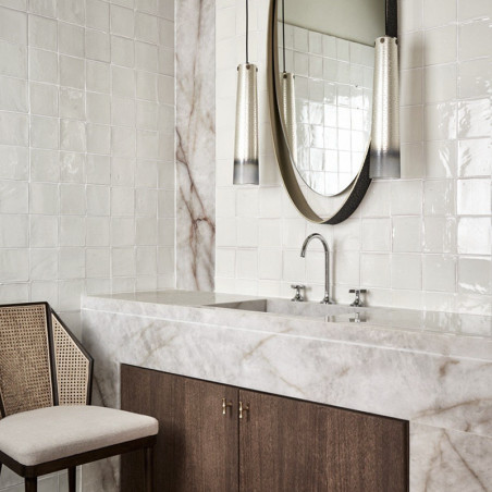 White terracotta tiles cover a bathroom wall, reflecting light. marble sink beneath, flanked by modern mirrors and lamps.