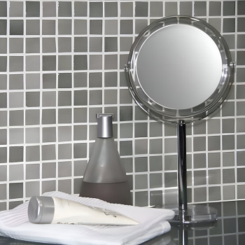 Mirror stands beside a pump bottle and cream tube on towels, reflecting against a grid-patterned glass mosaic wall.