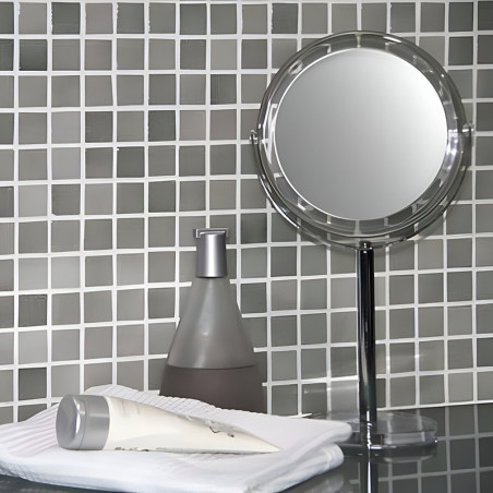 Mirror stands beside a pump bottle and cream tube on towels, reflecting against a grid-patterned glass mosaic wall.