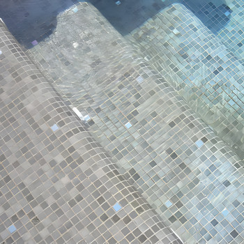 Glass tiles form steps submerged in water, creating a shimmering pattern under clear, gently moving pool water.