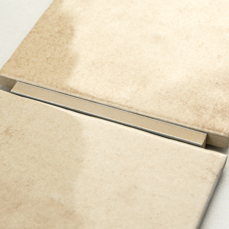 Twee beige keramische tegels rusten naast elkaar, gescheiden door een smalle metalen strip, op een gladde, neutraal gekleurde ac
