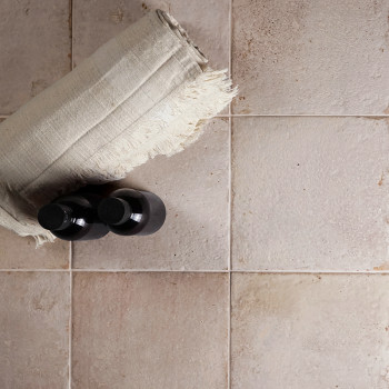 Una toalla de lino enrollada y dos botellas negras descansan sobre una cuadrícula de azulejos cuadrados de color beige, ligerame