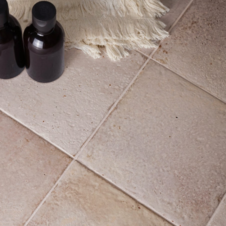 Deux bouteilles sombres et bouchées reposent sur des carreaux beiges aux surfaces texturées. Un tissu blanc cassé à franges se t