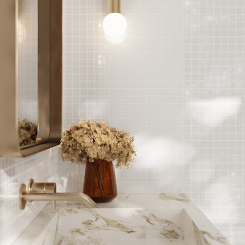 Un jarrón marrón sostiene flores secas, sobre un lavabo de mármol. Arriba, una bombilla encendida ilumina un baño alicatado.