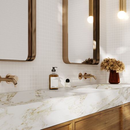 Los grifos de pared marrones se extienden sobre un lavabo de mármol blanco con vetas doradas. Arriba hay dos espejos, flanqueado