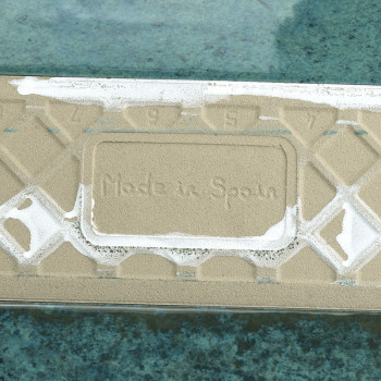 La losa rectangular, con la inscripción `Hecho en España`, yace plana, parcialmente cubierta de arena, contra una superficie de 