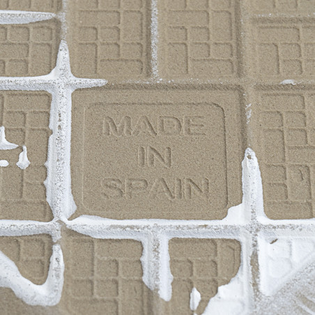 Un azulejo texturizado muestra el texto estampado `MADE IN SPAIN`, rodeado de líneas de lechada y parcialmente cubierto de lecha