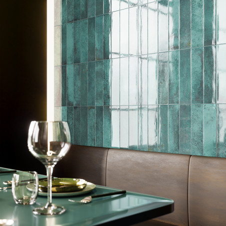 Los azulejos rectangulares de color verde azulado cubren una pared y reflejan la luz en un comedor. Un vaso y unos cubiertos des